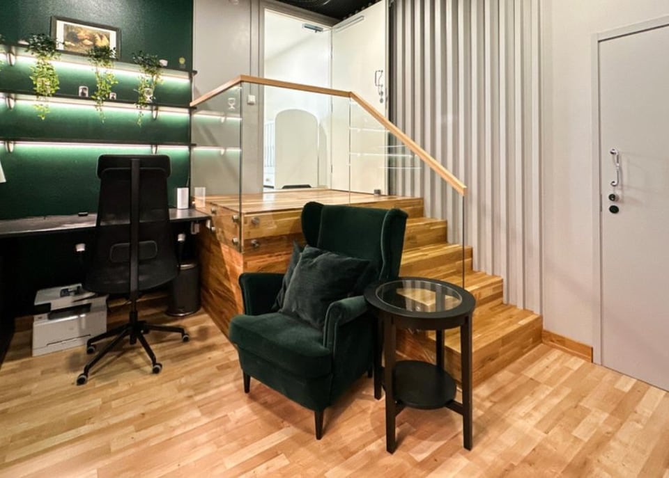 Green home office nook with a velvet wing armchair beside an open-tread oak staircase behind a glass balustrade