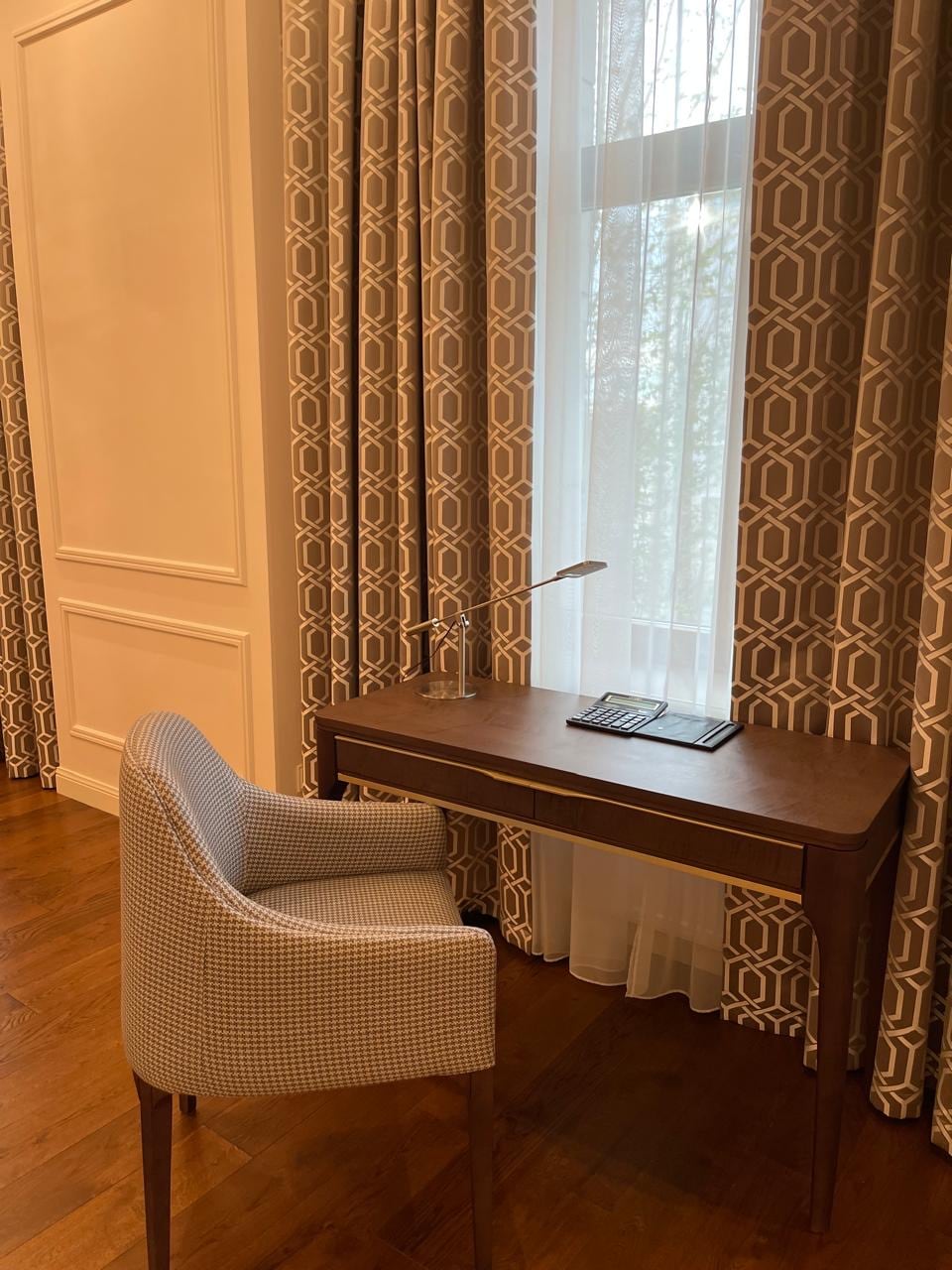 Writing desk against a patterned curtain, with a houndstooth armchair and an articulated desk lamp